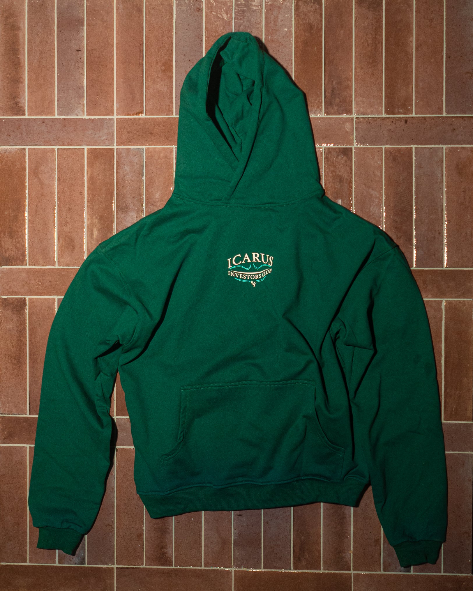 Green hoodie with a logo on the chest against a brown tiled wall